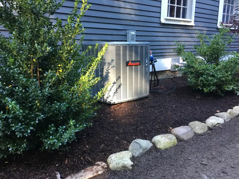 Amana central air conditioner condenser installed in a landscaped bed beside a clapboard home by A.J. LeBlanc Heating in Southern NH