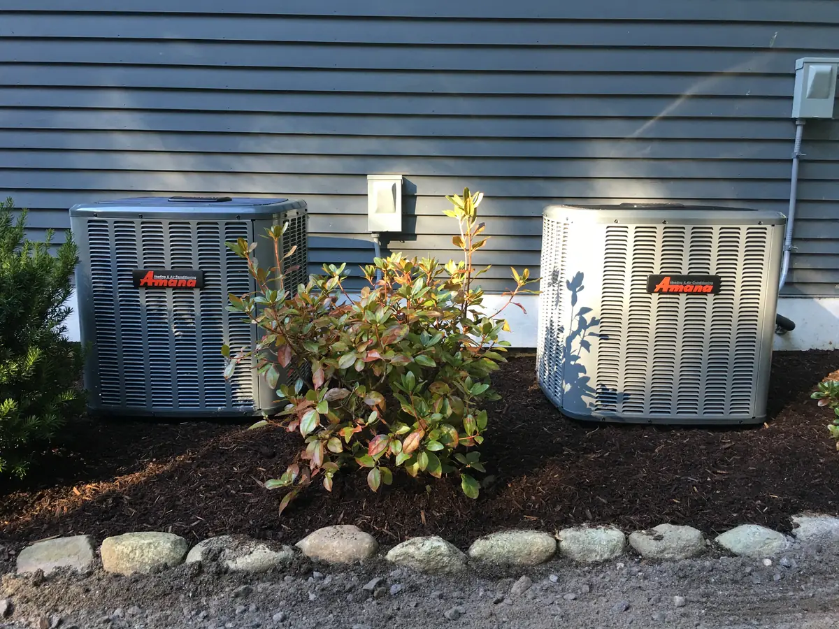 Dual Amana central air conditioners installed in a landscaped side yard by A.J. LeBlanc Heating in Southern NH