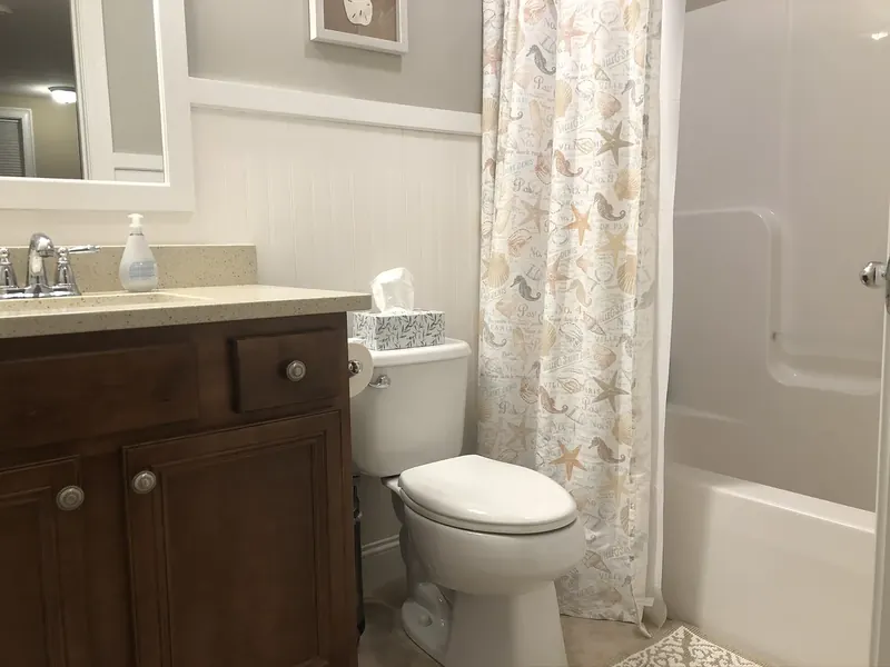 Coastal-themed bathroom with toilet, brown vanity with granite counter, white beadboard, and tub shower with seashell shower curtain installed by A.J. LeBlanc Heating in a Southern NH home