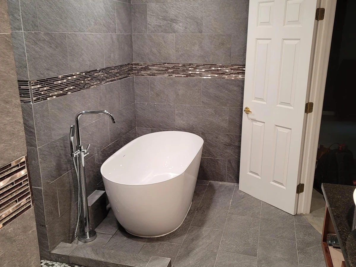 Modern freestanding oval bathtub with chrome floor-mount filler in a gray porcelain tile bathroom with glass mosaic accent band installed by A.J. LeBlanc Heating in Southern NH