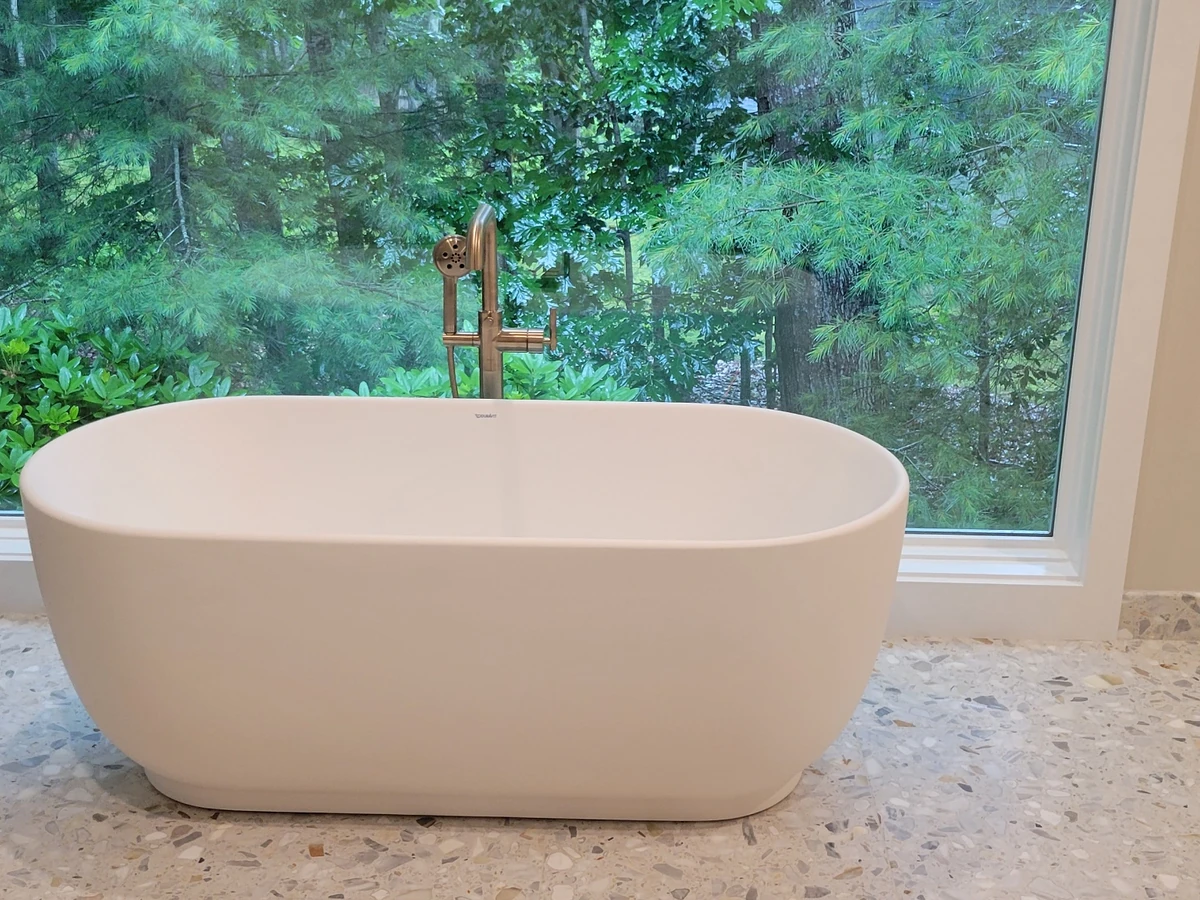 Freestanding bathtub with brushed brass floor-mount filler set in front of a large picture window overlooking the Southern NH forest, installed by A.J. LeBlanc Heating