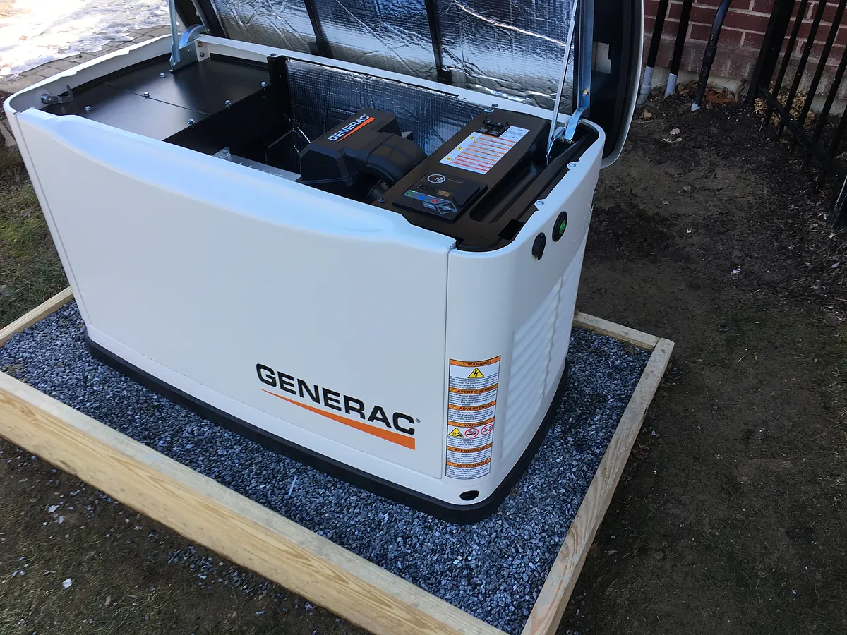 Generac whole-home standby generator with the hood open showing the internal battery and engine compartment during installation on a wood-framed gravel pad by A.J. LeBlanc Heating in Southern NH