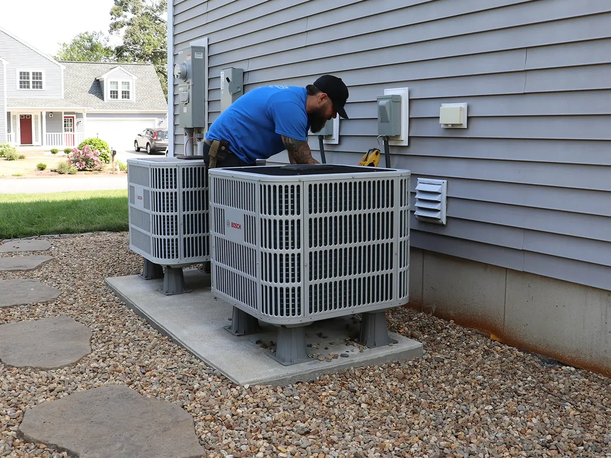 A.J. LeBlanc Heating technician servicing two outdoor Bosch central air conditioner condenser units on a gravel pad beside a Southern NH home