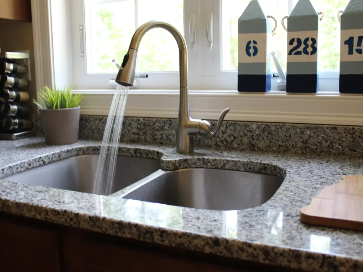 Chrome kitchen faucet running water into a stainless steel double-bowl undermount sink in a granite counter, installed by A.J. LeBlanc Heating in a Southern NH home