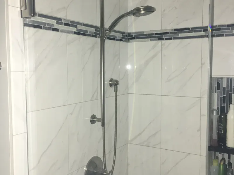 White marble tile shower with brushed nickel slide bar and handheld wand, glass mosaic accent band, and corner shelving installed by A.J. LeBlanc Heating in a Southern NH home