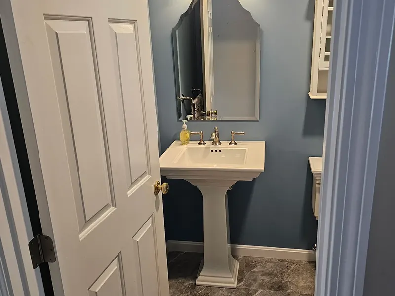 Pedestal sink with brushed nickel widespread faucet, arched mirror, two-light sconce, and marble tile floor installed by A.J. LeBlanc Heating in a Southern NH half bath