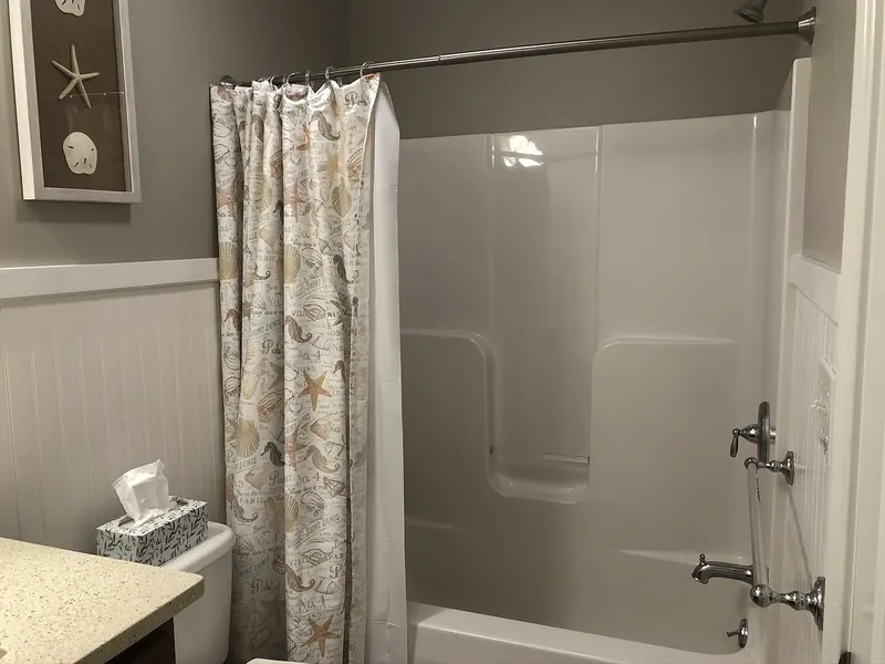 Tub and shower combo with one-piece fiberglass surround, chrome fixtures, beadboard wainscoting, and beach-themed shower curtain installed by A.J. LeBlanc Heating in a Southern NH home