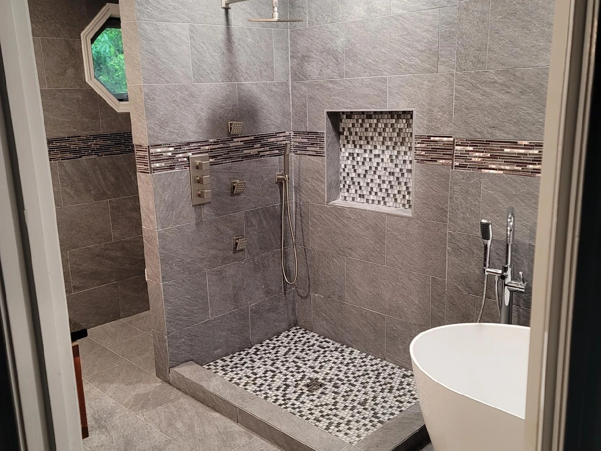 Walk-in shower with brushed nickel rain head, body sprays, handheld wand, recessed niche, pebble floor, and glass mosaic accent bands installed by A.J. LeBlanc Heating in a Southern NH home
